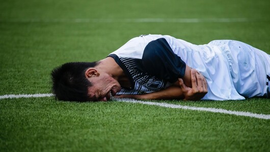 Bildet viser en fotballspiller som ligger på magen på en fotballbane. Spilleren har et fortvilt ansiktsuttrykk.