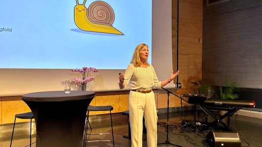 Hilde Inntjore står på scenen på likestillingskonferanse. På skjermen bak henne er det bilde av en tegnet snegle med et sneglehus med smilende munn og øyne.